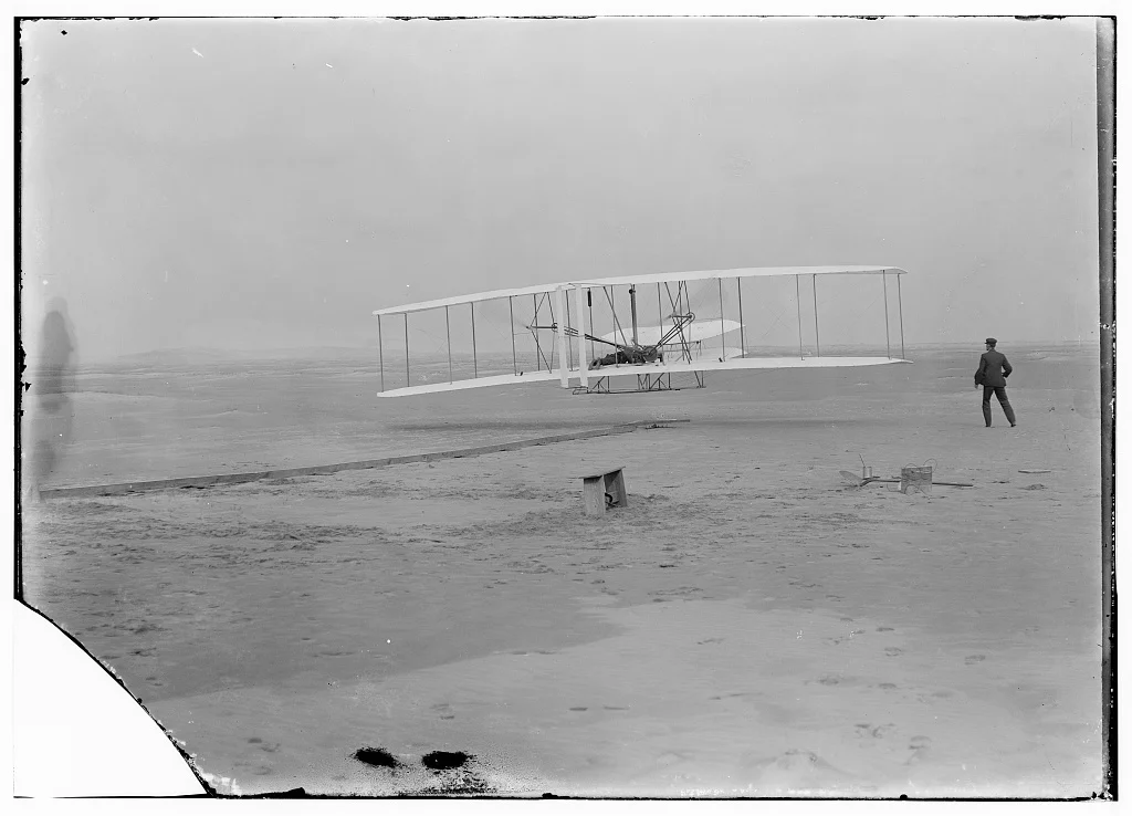 The Wright brothers’ first flight at Kitty Hawk, December 17, 1903, before restoration — the glass negative with its lower-left corner broken away and dark blotches along the bottom
